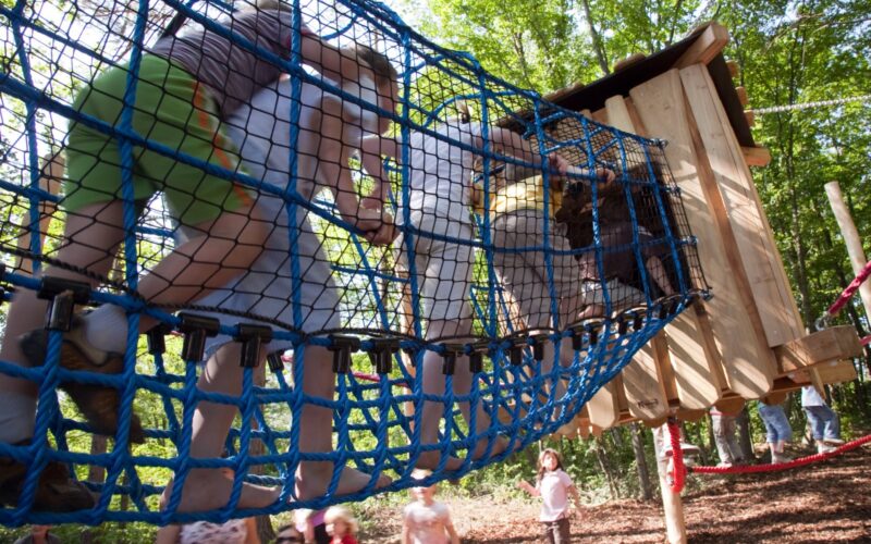 Kinder klettern über eine blaue Seilbrücke auf einem Außenspielplatz mit Bäumen und einer Holzkonstruktion im Hintergrund.