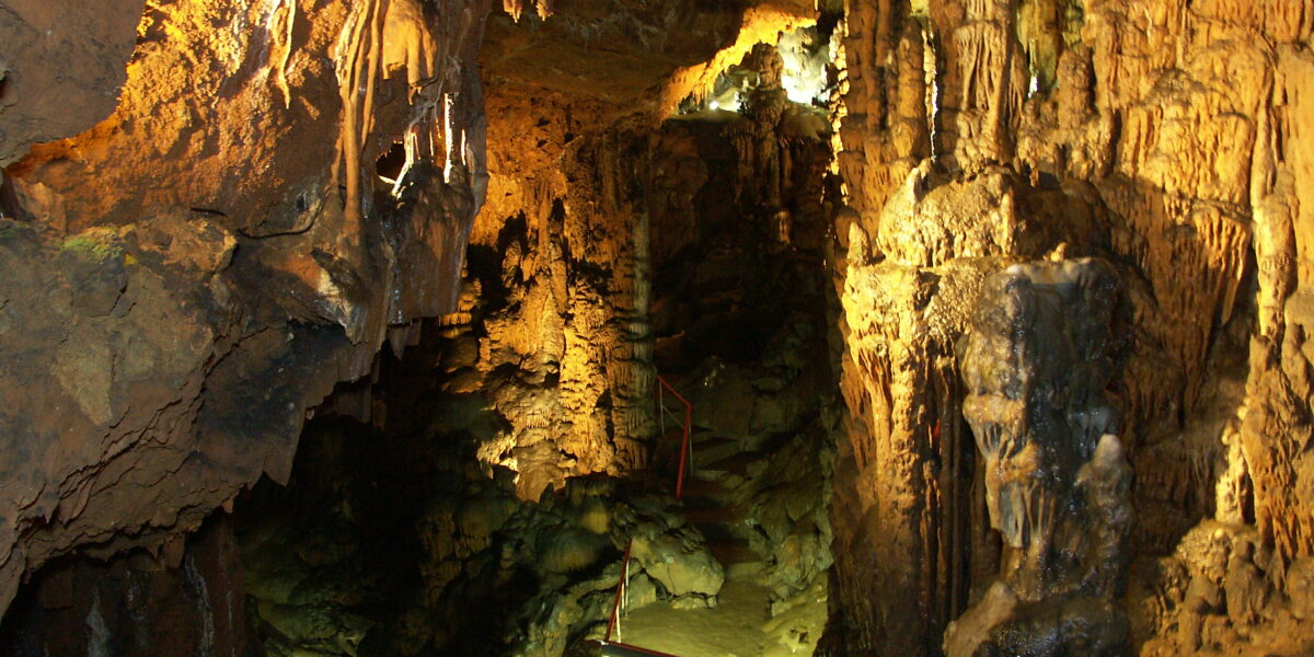Innenansicht einer Höhle mit Stalaktiten und Stalagmiten, die von künstlichen Lichtern beleuchtet werden, und einem Weg mit Geländern, der darunter sichtbar ist.