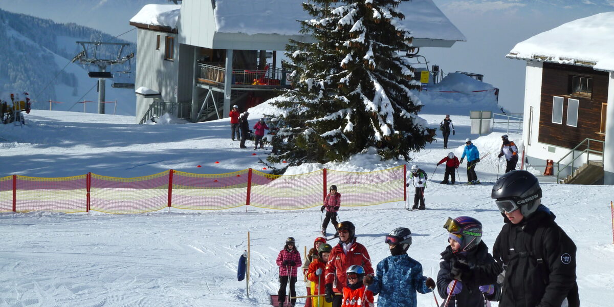 Kinder und Erwachsene in Skiausrüstung stehen in einem verschneiten Skigebiet auf einem Förderband in einer Reihe, im Hintergrund sind Berge, Bäume und Gebäude zu sehen.