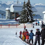 Kinder und Erwachsene in Skiausrüstung stehen in einem verschneiten Skigebiet auf einem Förderband in einer Reihe, im Hintergrund sind Berge, Bäume und Gebäude zu sehen.