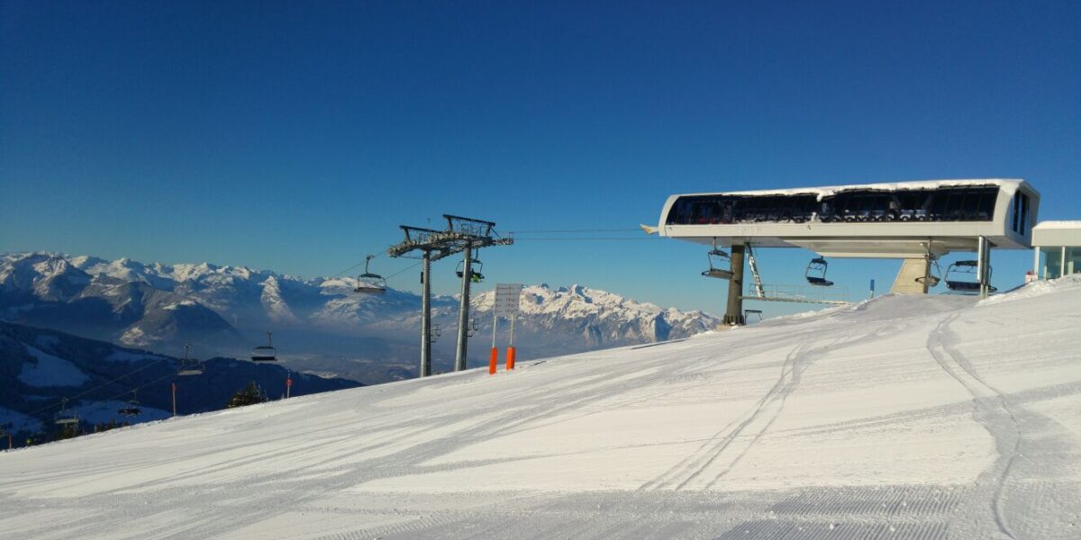Eine schneebedeckte Skipiste mit einer Skiliftstation, Skiliftsesseln und Bergketten im Hintergrund unter einem klaren blauen Himmel.