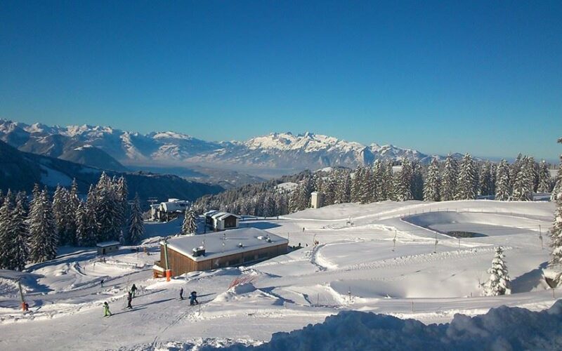 Schneebedecktes Skigebiet mit Skifahrern, umgeben von Kiefern und Bergen unter einem klaren blauen Himmel. Im Vordergrund sind Gebäude und Skispuren zu sehen.