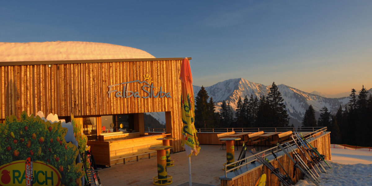 Eine hölzerne Skihütte mit Sitzgelegenheiten im Freien bietet Aussicht auf die schneebedeckten Berge bei Sonnenuntergang. Draußen steht die Skiausrüstung, und in der Nähe stehen Kiefern.