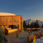 Eine hölzerne Skihütte mit Sitzgelegenheiten im Freien bietet Aussicht auf die schneebedeckten Berge bei Sonnenuntergang. Draußen steht die Skiausrüstung, und in der Nähe stehen Kiefern.
