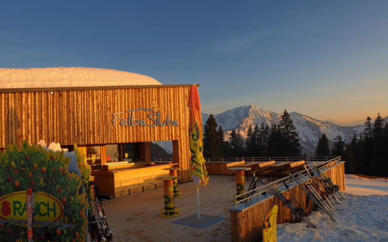 Eine hölzerne Skihütte mit Sitzgelegenheiten im Freien bietet Aussicht auf die schneebedeckten Berge bei Sonnenuntergang. Draußen steht die Skiausrüstung, und in der Nähe stehen Kiefern.