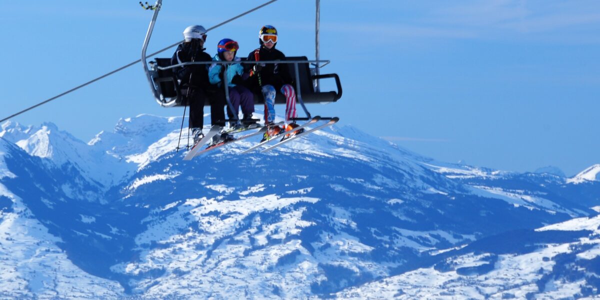 Drei Personen in Skiausrüstung fahren mit einem Sessellift über eine verschneite Berglandschaft mit klarem blauen Himmel im Hintergrund.