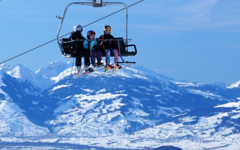 Drei Personen in Skiausrüstung fahren mit einem Sessellift über eine verschneite Berglandschaft mit klarem blauen Himmel im Hintergrund.