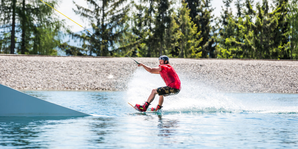 Eine Person, die ein rotes Hemd, einen Helm und kurze Hosen trägt, fährt mit einem Wakeboard auf einem Seilsystem über ein ruhiges Gewässer mit Bäumen im Hintergrund.
