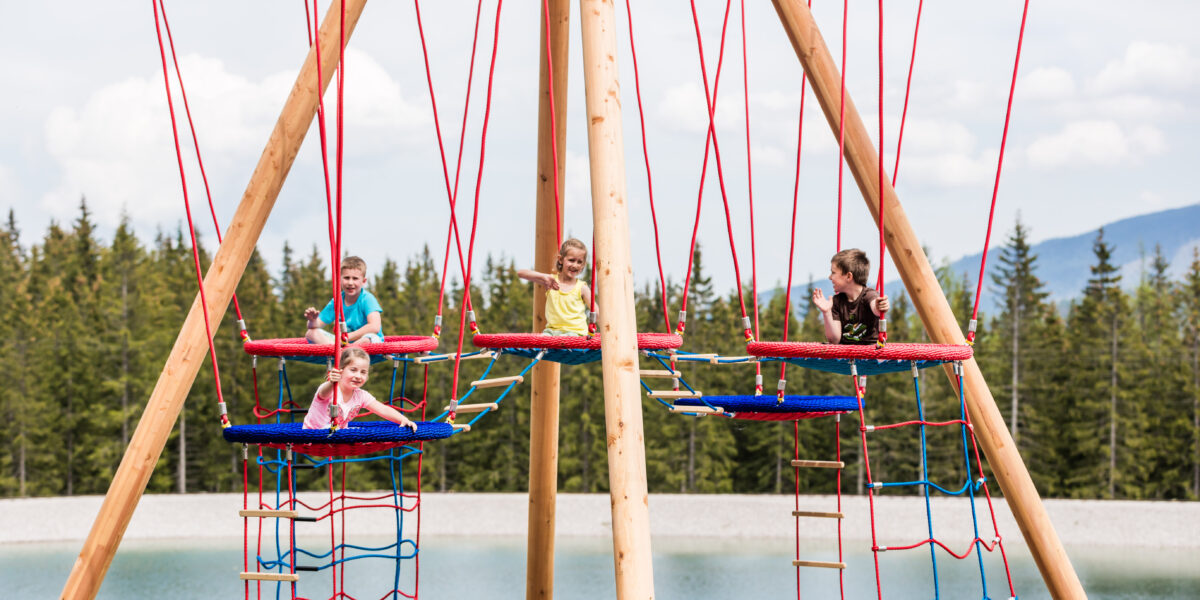 Vier Kinder sitzen auf aufgehängten Seilplattformen mit roten Schaukeln und Kletternetzen, mit einem See und Bäumen im Hintergrund an einem bewölkten Tag.