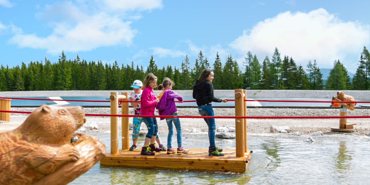 Vier Kinder stehen auf einem schwimmenden Holzfloß, das ein rotes Seil hält, und überqueren eine flache Wasserfläche mit Bäumen und blauem Himmel im Hintergrund.