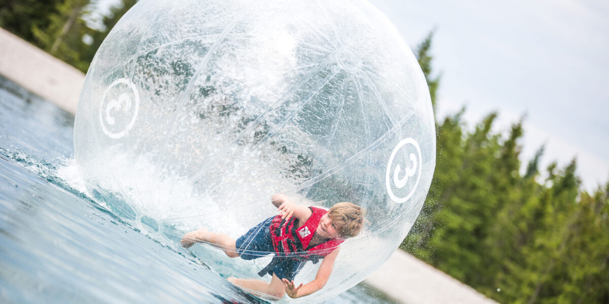 Ein Kind in einer Schwimmweste bewegt sich in einer großen aufblasbaren Plastikkugel, die auf dem Wasser schwimmt, mit Bäumen im Hintergrund.