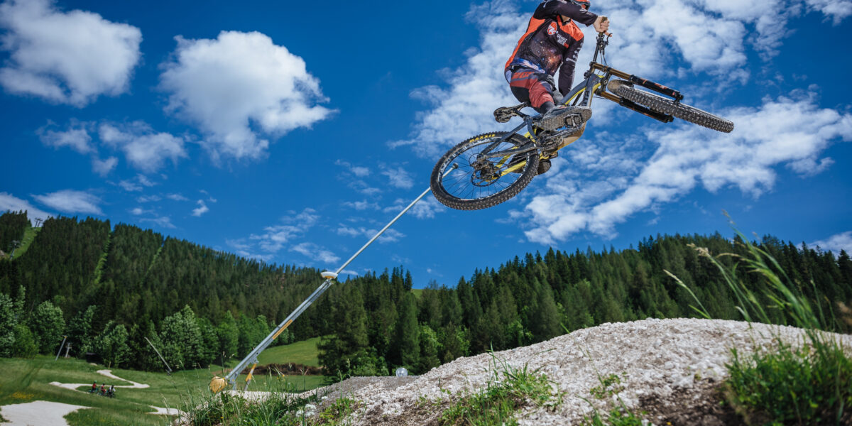 Ein Mountainbiker in Schutzkleidung springt hoch von einer unbefestigten Rampe in einem grasbewachsenen Waldgebiet unter blauem Himmel mit vereinzelten Wolken.