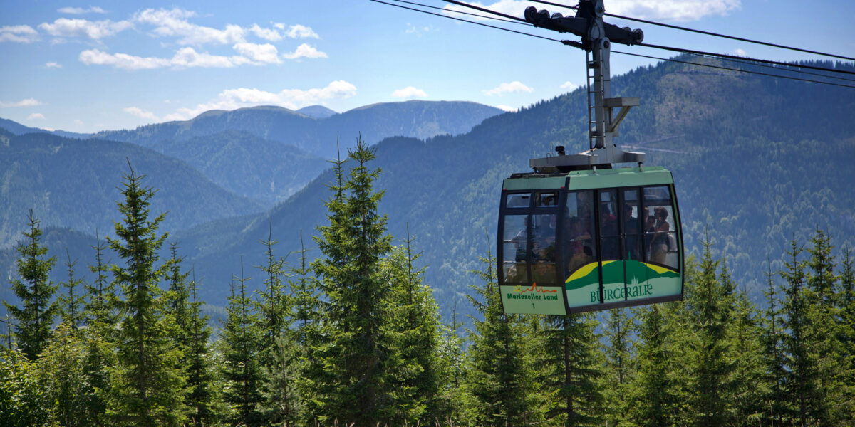 Eine grüne Seilbahn fährt über Pinienbäume, im Hintergrund sind Bergketten zu sehen, und der Himmel ist teilweise bewölkt.