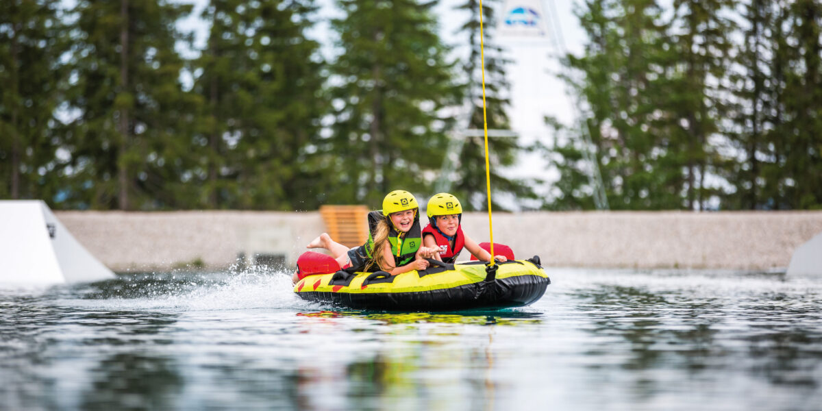Zwei Personen mit Helmen fahren auf einem aufblasbaren Wasserschlauch, lächeln und halten sich fest, während sie über ein Gewässer mit Bäumen im Hintergrund gezogen werden.