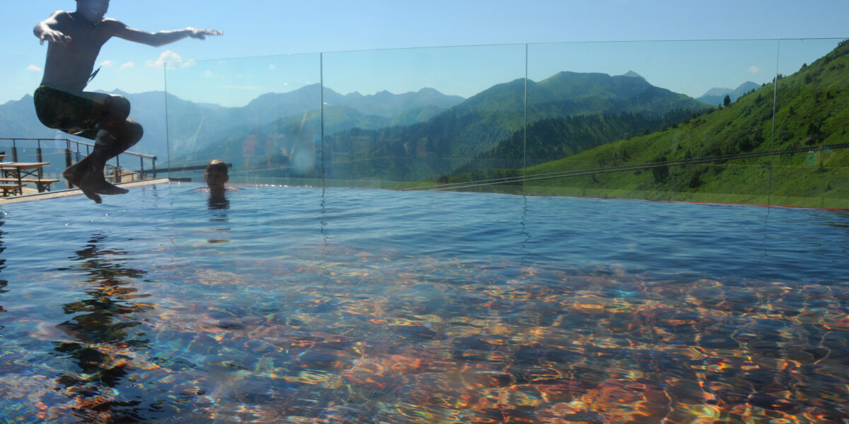 Eine Person springt an einem sonnigen Tag in ein Freibad mit Bergen im Hintergrund. Eine andere Person befindet sich im Wasser in der Nähe des Beckenrandes.