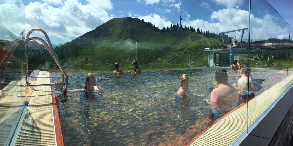 Die Menschen schwimmen und entspannen sich in einem Freibad mit Glaswänden, mit Blick auf grüne Berge und eine Seilbahnstation unter einem teilweise bewölkten Himmel.