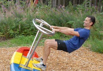 Ein Mann in Sportkleidung hält sich an einem Metallrad fest, das an einem farbenfrohen Spielplatzgerät im Freien befestigt ist, lehnt sich zurück und lächelt. Im Hintergrund sind Bäume und Grünflächen zu sehen.