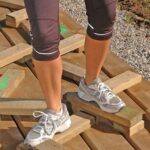 Eine Person, die Sportschuhe und Capri-Leggins trägt, steht im Freien auf unebenen Holzbrettern, in deren Nähe sich Kies und Erde befinden.
