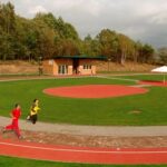 Zwei Personen joggen auf einer Laufbahn im Freien neben einem grasbewachsenen Feld mit roten Sportflächen, Bäumen und einem kleinen Gebäude im Hintergrund.