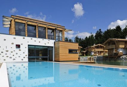 Modernes Gebäude mit großen Fenstern und Holzakzenten neben einem Außenpool, mit Bäumen und Häusern im Chalet-Stil im Hintergrund unter einem teilweise bewölkten Himmel.