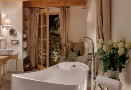 Eine moderne weiße Badewanne steht im Mittelpunkt eines gemütlichen Badezimmers mit Holzakzenten, einem Sessel, einem Strauß weißer Rosen und Glastüren nach draußen.