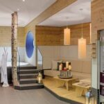 Moderne Spa-Lounge mit Holzakzenten, Birkendekor, einem Sitzbereich mit Kerzen, einer Wanduhr und einem Whirlpool, der teilweise im Hintergrund zu sehen ist.