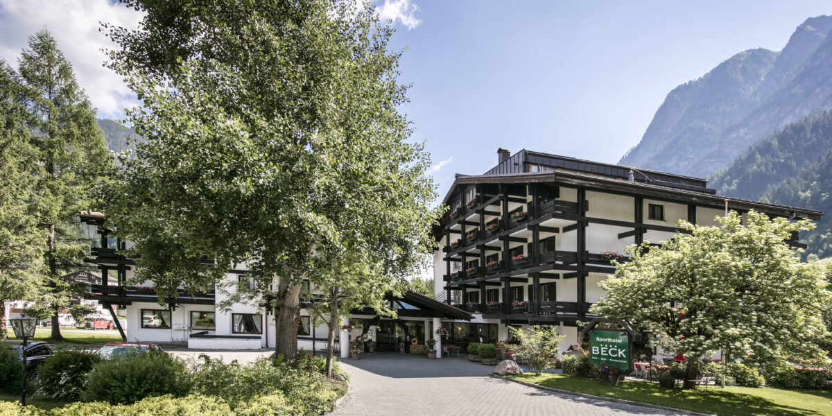 Ein mehrstöckiges Alpenhotel mit dunklen Holzbalkonen und weißen Wänden liegt zwischen Bäumen und Bergen unter blauem Himmel.