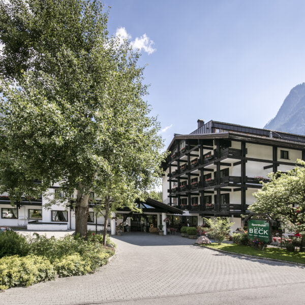 Ein mehrstöckiges Alpenhotel mit dunklen Holzbalkonen und weißen Wänden liegt zwischen Bäumen und Bergen unter blauem Himmel.