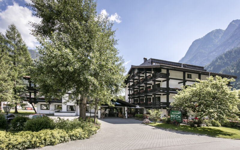 Ein mehrstöckiges Alpenhotel mit dunklen Holzbalkonen und weißen Wänden liegt zwischen Bäumen und Bergen unter blauem Himmel.