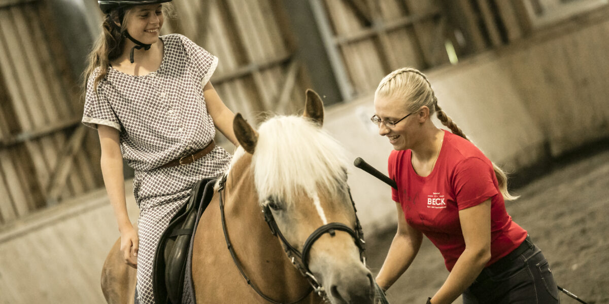 Ein junges Mädchen, das einen Helm trägt, sitzt auf einem Pferd, während ein Reitlehrer in einem roten Hemd in einer Reithalle daneben steht.