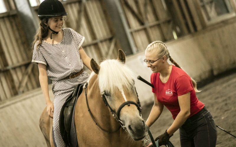Ein junges Mädchen, das einen Helm trägt, sitzt auf einem Pferd, während ein Reitlehrer in einem roten Hemd in einer Reithalle daneben steht.