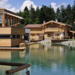 Holzhäuser im Chalet-Stil und ein modernes Gebäude umgeben einen klaren Teich mit einem bewaldeten Gebiet und blauem Himmel im Hintergrund.