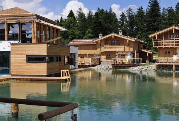 Holzhäuser im Chalet-Stil und ein modernes Gebäude umgeben einen klaren Teich mit einem bewaldeten Gebiet und blauem Himmel im Hintergrund.