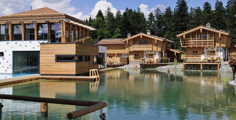 Holzhäuser im Chalet-Stil und ein modernes Gebäude umgeben einen klaren Teich mit einem bewaldeten Gebiet und blauem Himmel im Hintergrund.