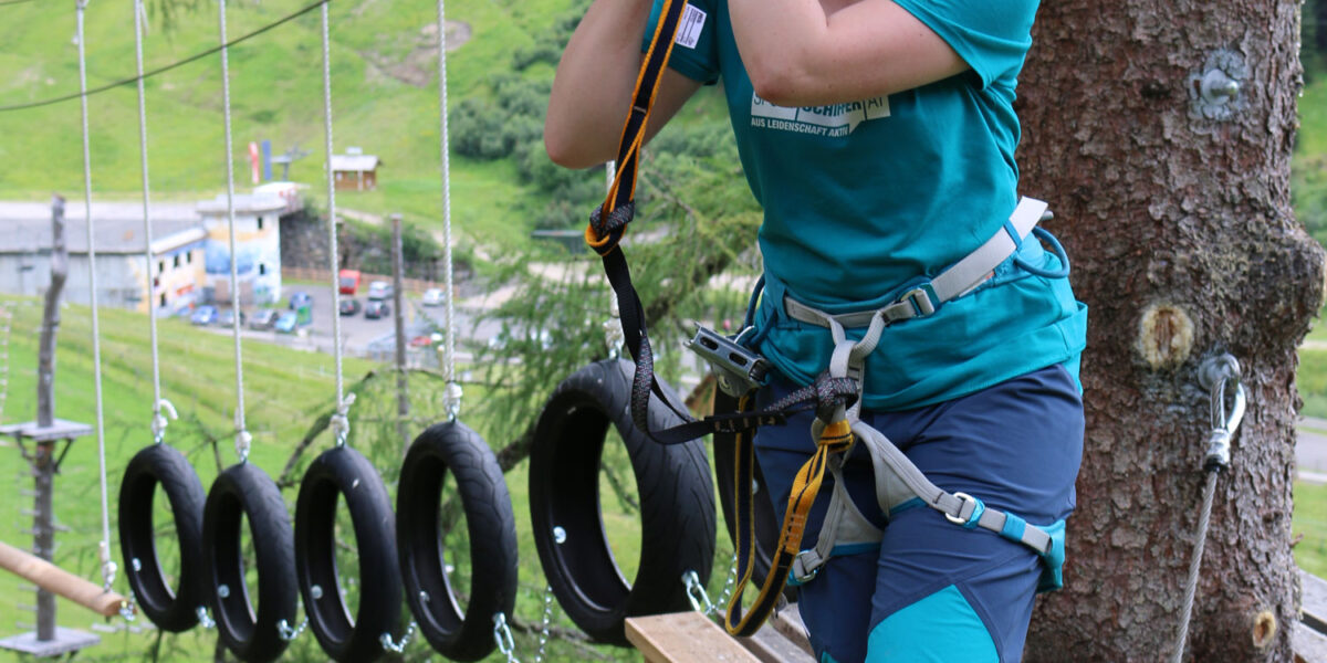 Eine Person mit Sicherheitsausrüstung steht auf einer Holzplattform in einem Hochseilgarten und hält sich an einem Seil fest, das an einem Gurt befestigt ist.