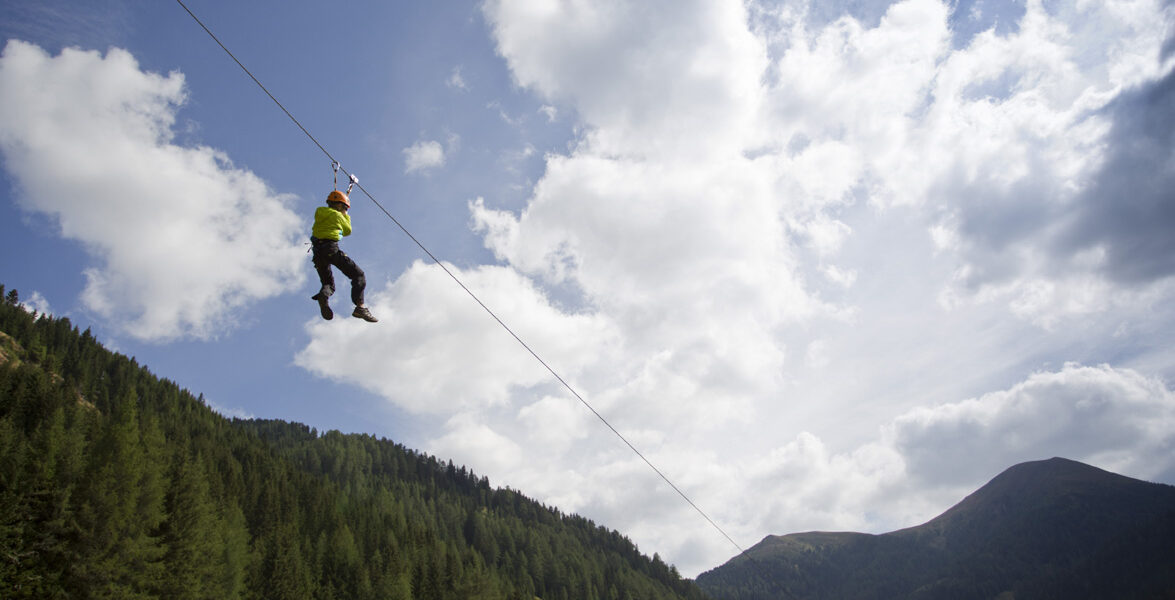 Eine Person, die einen Helm und ein Gurtzeug trägt, fährt auf einer Seilrutsche hoch über einem bewaldeten Tal mit Bergen und einem teilweise bewölkten Himmel im Hintergrund.