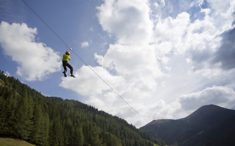 Eine Person, die einen Helm und ein Gurtzeug trägt, fährt auf einer Seilrutsche hoch über einem bewaldeten Tal mit Bergen und einem teilweise bewölkten Himmel im Hintergrund.