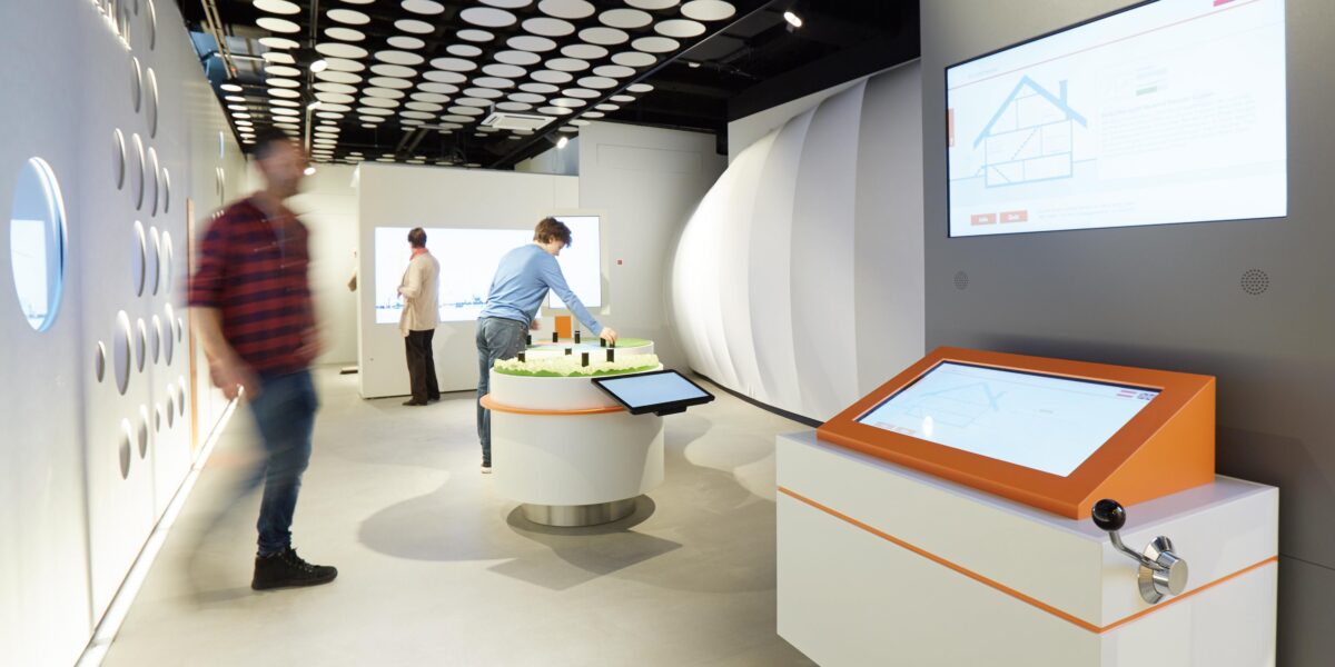 Mehrere Personen interagieren mit Touchscreen-Displays und einem Modell in einem modernen, hell erleuchteten Ausstellungsraum mit kreisförmigen Deckenplatten.