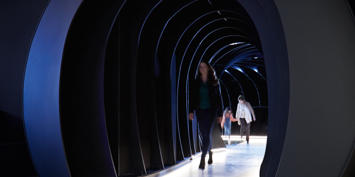 Man geht durch einen Tunnel mit geschwungenen, beleuchteten schwarzen Bögen und einem glatten Boden, der eine moderne und futuristische Atmosphäre schafft.