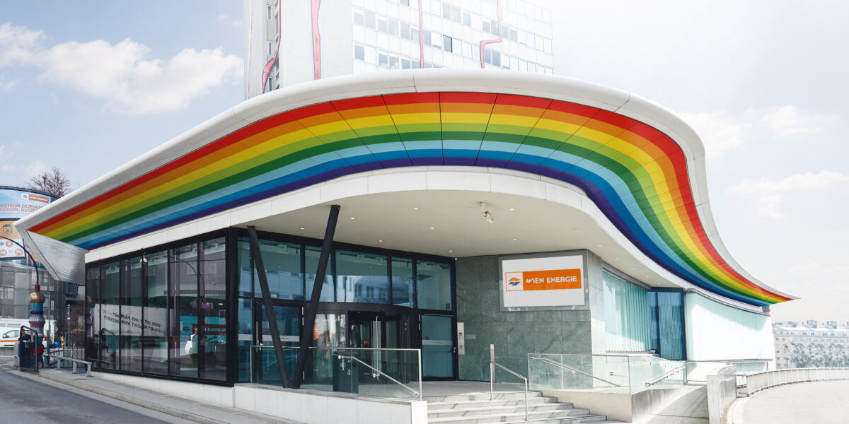 Modernes Gebäude mit großen Glasfenstern und einem markanten, wellenförmigen Dach mit einem regenbogenfarbenen Streifen entlang der Kante; Schild "Wien Energie" über dem Eingang.
