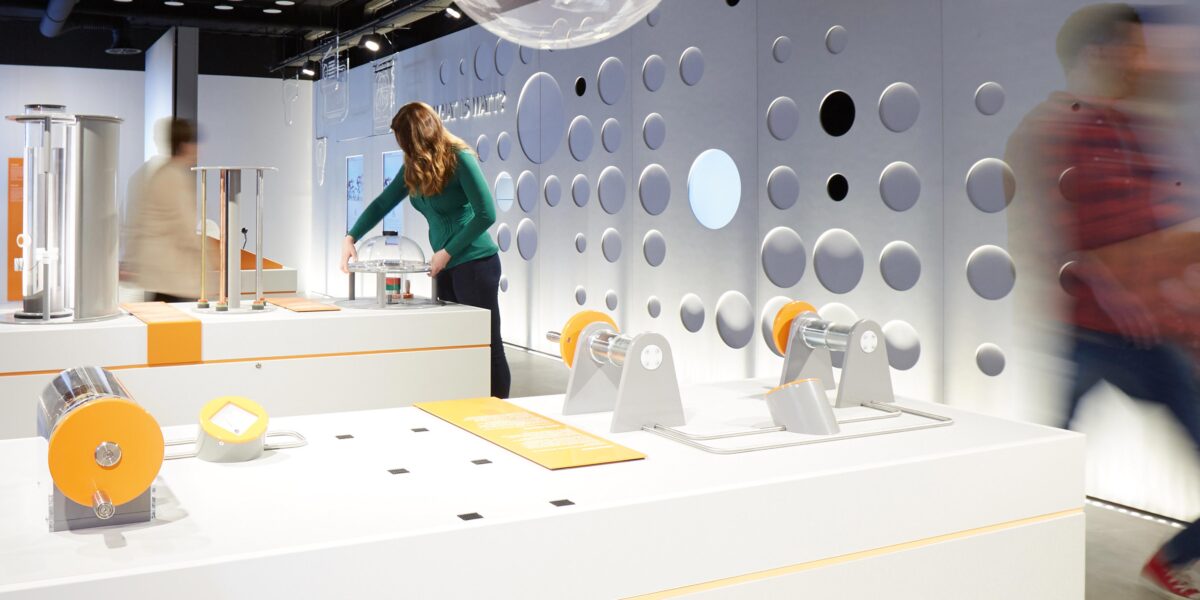 Eine Frau interagiert mit einem Exponat in einem modernen Wissenschaftsmuseum mit weißen Wänden, kreisförmigen Ausschnitten und wissenschaftlichen Displays; zwei verschwommene Besucher gehen vorbei.