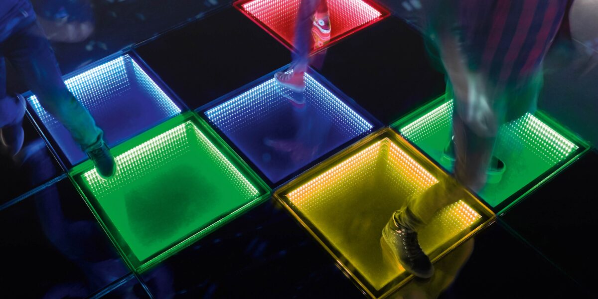 Menschen, die auf einem Glasboden mit beleuchteten Quadraten in Blau, Grün, Gelb und Rot auf einem dunklen Hintergrund gehen.