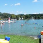 Menschen schwimmen und entspannen sich an einem sonnigen Tag an einem Strand am See, mit Segelbooten im Hintergrund und Schlauchbooten auf dem Rasen in der Nähe des Wassers.
