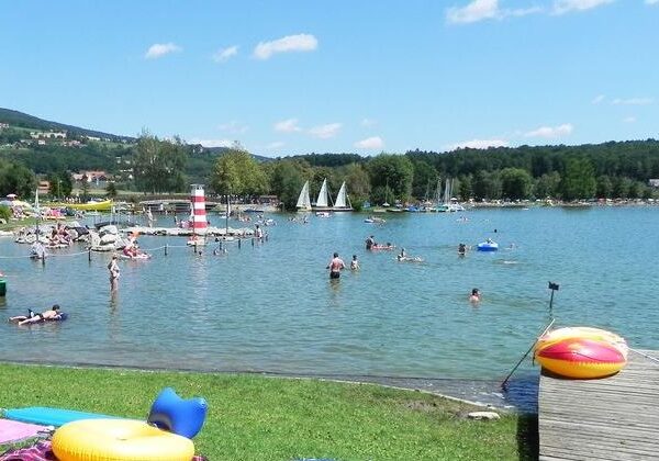 Menschen schwimmen und entspannen sich an einem sonnigen Tag an einem Strand am See, mit Segelbooten im Hintergrund und Schlauchbooten auf dem Rasen in der Nähe des Wassers.