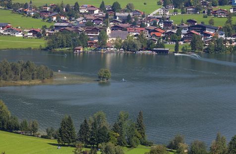 Eine kleine Stadt mit roten Dächern und einer Kirche liegt am Rande eines Sees, umgeben von grünen Feldern und Bäumen.