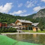 Holzgebäude mit Sitzgelegenheiten im Freien neben einem grün gefärbten Teich, umgeben von Grasflächen, Bäumen und Bergen unter einem teilweise bewölkten Himmel.