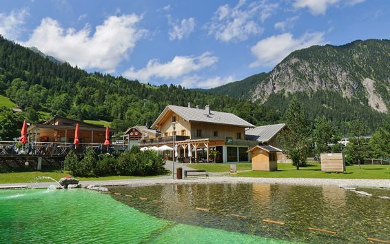 Holzgebäude mit Sitzgelegenheiten im Freien neben einem grün gefärbten Teich, umgeben von Grasflächen, Bäumen und Bergen unter einem teilweise bewölkten Himmel.