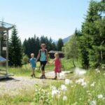 Ein Erwachsener und zwei Kinder gehen auf einem Pfad durch ein grasbewachsenes, bewaldetes Gebiet, vorbei an einem Informationsschild unter einem klaren Himmel.