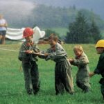 Vier Kinder mit Helmen und Overalls spritzen im Freien mit einem Schlauch Wasser, im Hintergrund sind zwei Erwachsene und eine Hügellandschaft zu sehen.