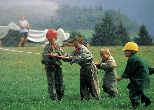 Vier Kinder mit Helmen und Overalls spritzen im Freien mit einem Schlauch Wasser, im Hintergrund sind zwei Erwachsene und eine Hügellandschaft zu sehen.